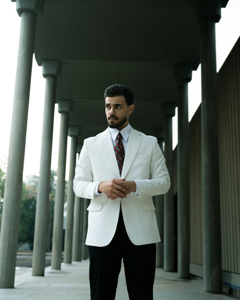 Confident man in a white suit standing between tall columns outdoors.